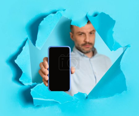 Photo for Middle aged man showing smartphone with blank empty screen through torn in blue paper wall background, recommending great online offer, app or website, mockup, collage - Royalty Free Image