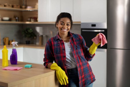 Téléchargez les photos : Heureuse femme afro-américaine millénaire en gants en caoutchouc avec chiffon près de la table profiter des tâches ménagères dans l'intérieur de la cuisine minimaliste. Service de nettoyage, mode de vie, hygiène à la maison en raison de l'épidémie de covidé-19 - en image libre de droit