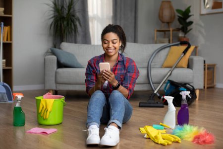 Photo for App for social networks and communication. Happy millennial black lady in apron sits on floor with cleaning supplies chatting on phone in room interior. Rest from household chores with device at home - Royalty Free Image