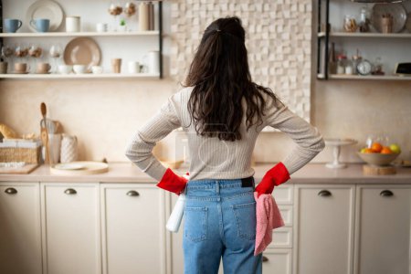 Photo for Busy young woman in rubber gloves with spray and rag in hands looking at cleaning result in light kitchen interior, back view, copy space. Household chores, cleanliness and hygiene - Royalty Free Image