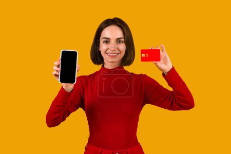 Foto de Sonriente mujer bastante joven en rojo celebración de teléfono inteligente moderno con pantalla en blanco y tarjeta de crédito aislado en fondo de estudio naranja, pagar por bienes y servicios en Internet, compras en línea, maqueta - Imagen libre de derechos