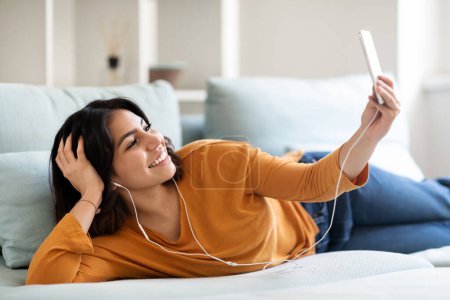 Téléchargez les photos : Femme arabe souriante avec smartphone et écouteurs se détendre sur le canapé à la maison, Joyeux jeune femme du Moyen-Orient écoutant de la musique en ligne ou faisant un appel vidéo sur téléphone mobile, Espace libre - en image libre de droit