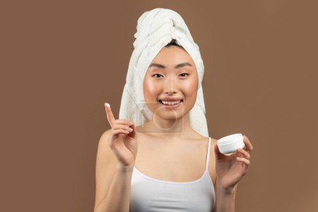 Téléchargez les photos : Une routine de soins. Belle dame asiatique tenant pot avec crème hydratante et la peau nourrissante tout en se tenant sur fond de studio brun - en image libre de droit