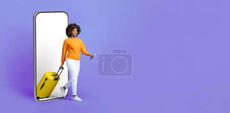Téléchargez les photos : Cool jolie jeune voyageuse noire marchant depuis un grand téléphone portable avec écran blanc vide, portant des bagages jaunes, tenant un passeport avec des billets, maquette, fond de studio violet, espace de copie - en image libre de droit