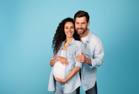 Photo for Smiling young european male hugging pregnant female in casual with big belly, isolated on blue background, studio. Family love and relationship, baby expectation, happy future parents and lifestyle - Royalty Free Image