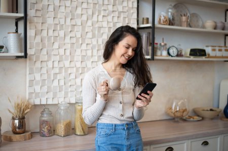 Foto de Mujer joven feliz usando teléfono inteligente mientras bebe té de la mañana o café en el interior de la cocina en casa, espacio libre. Emocionado señora leyendo mensajes de texto en su teléfono celular y sonriendo - Imagen libre de derechos