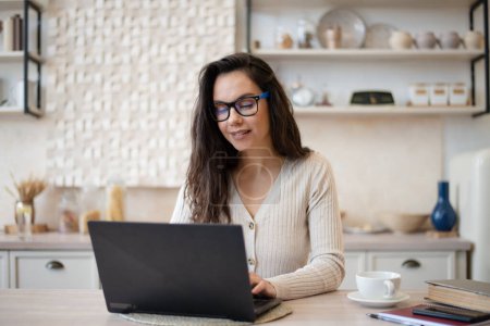Téléchargez les photos : Joyeux jeune femme pigiste travaillant de la maison, assis à table et utilisant un ordinateur portable, dactylographiant un e-mail ou surfant sur Internet, intérieur de la cuisine, espace libre - en image libre de droit