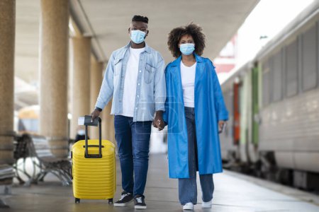 Téléchargez les photos : African American Couple Wearing Protective Medical Masks Walking At Railway Station, Young Black Man And Woman Carrying Luggage While Passing Platform, Holding Hands And Looking To Camera - en image libre de droit