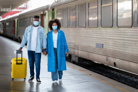 Foto de Young Black Couple Wearing Protective Medical Masks Walking With Suitcase At Railway Station, African American Spouses Holding Hands While Going To Train, Travelling During Pandemic, Copy Space - Imagen libre de derechos