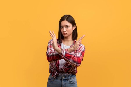 Foto de Portrait of assertive asian lady showing STOP or ENOUGH gesture on yellow studio background. Young korean female rejecting something, expressing her denial, saying NO - Imagen libre de derechos