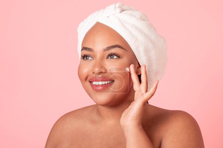 Téléchargez les photos : Skin care concept. Closeup portrait of black plus size lady applying moisturizing cream on face, posing over pink studio background and smiling - en image libre de droit