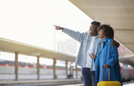 Téléchargez les photos : Happy African American Couple With Luggage Standing On Platform At Railway Station, Cheerful Young Black Spouses Waiting For Train Together, Woman Holding Smartphone And Man Pointing Away, Copy Space - en image libre de droit
