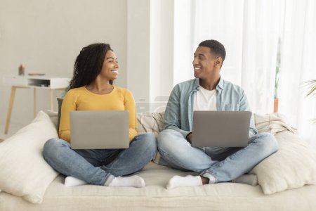 Téléchargez les photos : Smiling millennial african american male and female with laptops looking at each other in living room interior. Work, business and freelance, social distancing and new normal, technology at home - en image libre de droit