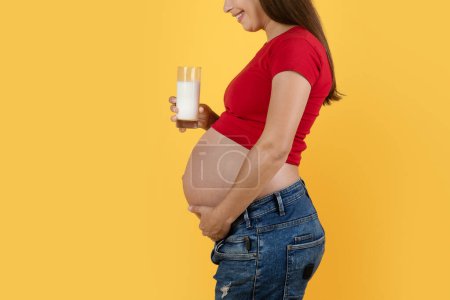 Téléchargez les photos : Pregnancy And Dairy Foods. Side View Of Pregnant Female Holding Glass Of Milk While Standing Isolated On Yellow Background, Young Expectant Woman Caressing Her Belly And Enjoying Healthy Drink - en image libre de droit