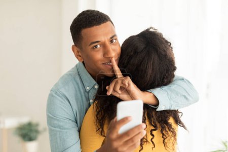 Téléchargez les photos : Cheerful millennial african american husband hug wife show silence sign, hold smartphone in living room interior, free space. Secret shh gesture, online surprise, app for gift, relationship at home - en image libre de droit