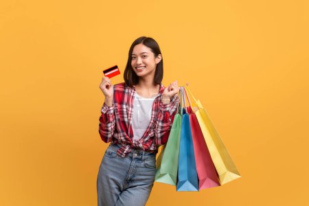 Téléchargez les photos : Cashback concept. Happy asian lady posing with credit card and colorful shopping bags over yellow studio background. Excited woman enjoying big discounts and offers, copy space - en image libre de droit