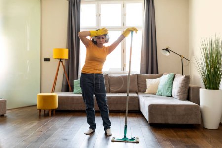 Téléchargez les photos : Young Arab Woman Feeling Tired While Making Cleaning In Living Room, Middle Eastern Female Wearing Headphones Exhausted After Mopping Floor At Home, Having Break And Wiping Forehead, Free Space - en image libre de droit