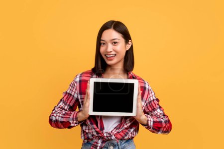Téléchargez les photos : Smiling asian lady showing digital tablet blank screen, advertising app or website, standing over yellow studio background. Woman holding device with empty monitor. Mockup - en image libre de droit