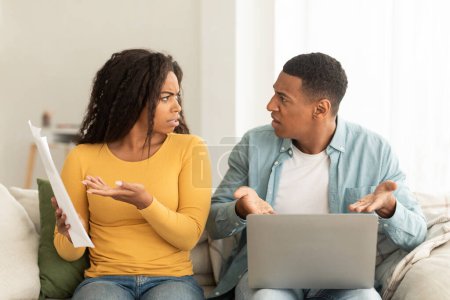 Foto de Angry sad millennial african american couple quarrel over debts and bills with papers and laptop in living room interior. People emotions, problems with finances, bankruptcy, stress and crisis at home - Imagen libre de derechos