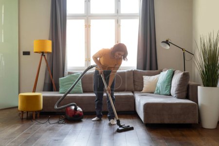 Téléchargez les photos : Young Arab Woman Using Vacuum Cleaner While Tidying Living Room, Middle Eastern Housewife Cleaning Floor At Home With Hoover, Millennial Female Enjoying Making Domestic Chores, Copy Space - en image libre de droit