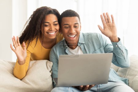 Photo for Technology, meeting remotely. Glad millennial african american couple looking at laptop, have video call, waving hands in light living room interior. Greeting, say hi, hello and communication at home - Royalty Free Image