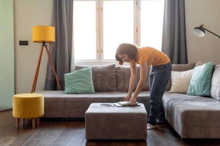 Photo for Smiling Middle Eastern Female Tidying Stylish Living Room At Home, Young Arab Woman Making Spring Cleaning, Millennial Housewife Placing Belongings In Order, Making Domestic Chores, Copy Space - Royalty Free Image
