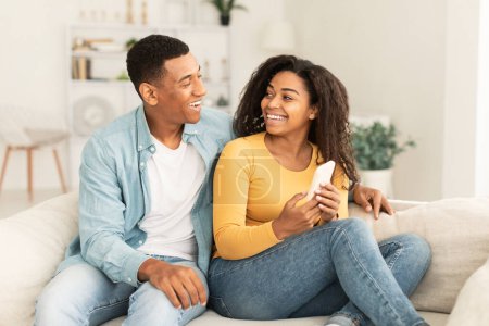 Photo for Cheerful millennial african american wife showing smartphone to husband, recommend new app in light living room interior. Technology and social networks for communication, online ad and offer at home - Royalty Free Image