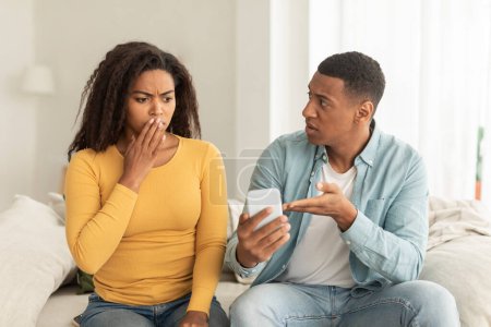 Photo for Sad millennial african american husband with smartphone scolding offended confused wife in light living room interior. Addiction to gadget and social networks, quarrel, relationship problems at home - Royalty Free Image