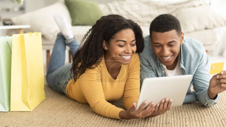 Cheerful millennial african american couple with bags with purchases lying on floor with tablet, credit card, shopping online in living room interior, panorama. Sale for shopaholics, delivery at home
