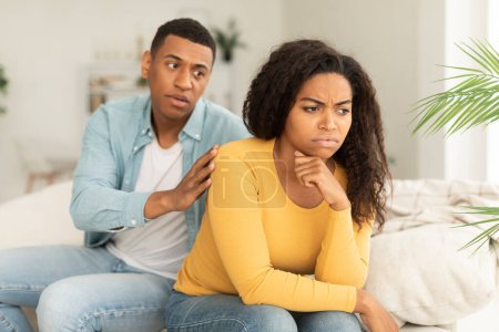 Photo for Sad husband calms offended upset millennial african american wife, ignore guy after quarrel in living room interior. Forgiveness, sorry, caring, support and emotions, relationships problems at home - Royalty Free Image