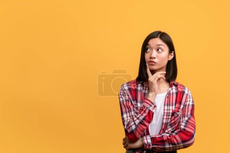 Photo for Let me think. Pensive asian lady looking at copy space, thinking about offer or deal and touching chin, standing over yellow studio background, banner - Royalty Free Image