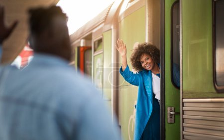 Foto de Happy Black Woman Waving Hand To Boyfriend While Standing In Train Door At Railway Station, Joyful Beautiful African American Female Greeting Her Spouse After Arrival Or Saying Bye Before Leaving - Imagen libre de derechos