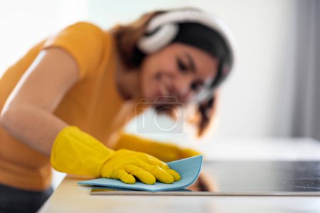 Foto de Smiling Young Woman Cleaning Induction Cooktop Surface With Rag In Kitchen, Happy Arab Female Wearing Rubber Gloves And Headphones Making Domestic Chores, Enjoying Tidying Home, Closeup - Imagen libre de derechos