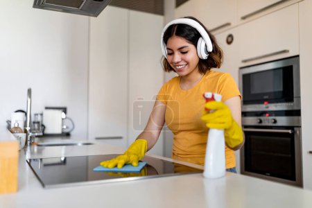Photo for Happy Young Arab Lady Wearing Headphones Doing Cleaning In Kitchen At Home, Smiling Middle Eastern Woman Washing Induction Cooktop Surface With Rag And Listening Favorite Music, Free Space - Royalty Free Image