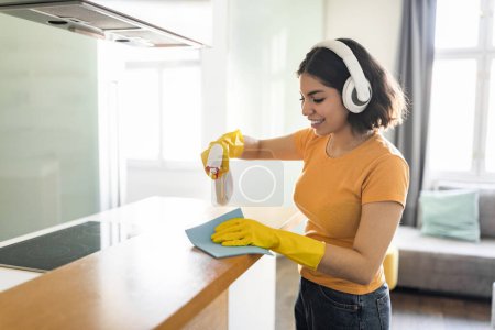 Foto de Domestic Chores Concept. Smiling Arab Woman Doing Cleaning In Kitchen, Happy Young Middle Eastern Female Wearing Wireless Headphones Using Detergent Sprayer And Rag While Tidying Home, Free Space - Imagen libre de derechos