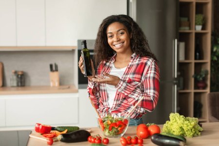 Téléchargez les photos : Smiling millennial black lady preparing salad, show oil in kitchen interior with organic vegetables, free space. Recommendation, offer and ad, food blog, health care, vegan food, cooking lunch at home - en image libre de droit