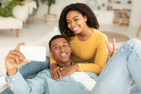 Photo for Smiling millennial african american wife hugging husband, relaxing on sofa and taking selfie on smartphone in living room interior. Love, romance and technology, app for photo, video call at home - Royalty Free Image