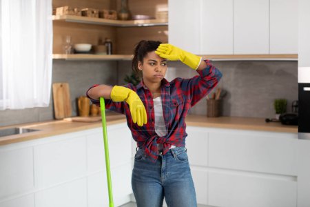 Téléchargez les photos : Unhappy millennial african american housewife in rubber gloves wipes her forehead and mop floor in minimalist kitchen interior. Tired of cleaning house alone, a lot of work on weekends and service - en image libre de droit