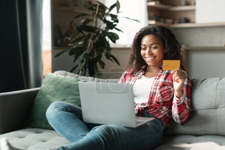 Foto de Online shopping, big sale. Smiling millennial black lady shopaholic use computer and credit card to check banking account, sit on sofa in interior living room. Cashback, finance and lifestyle at home - Imagen libre de derechos