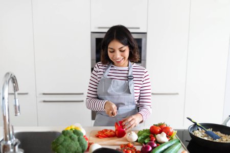 Foto de Healthy diet, cooking at home concept. Cheerful attractive young middle eastern lady wearing apron preparing delicious meal at kitchen, cutting organic vegetables and smiling, copy space - Imagen libre de derechos