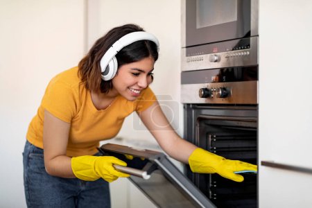 Photo for Happy Young Arab Woman In Headphones Wiping Oven While Cleaning In Kitchen, Cheerful Middle Eastern Housewife Wearing Rubber Gloves Tidying Home And Listening Favorite Music, Closeup Shot - Royalty Free Image