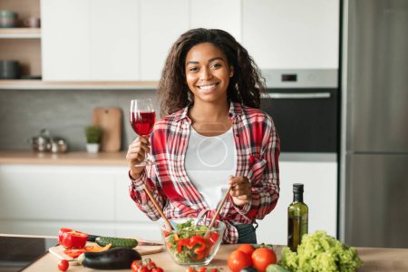 Photo for Smiling pretty millennial black female preparing salad, hold wine glass at table with organic vegetables in kitchen interior. Holiday celebration, rest and relax at night, date and lifestyle at home - Royalty Free Image