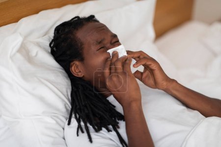 Téléchargez les photos : Sick African American Man Blowing Nose In Paper Tissue While Lying In Bed At Home, Young Black Male Feeling Unwell While Having Rhinitis Or Allergy, Suffering Seasonal Flu Or Cold, Closeup Shot - en image libre de droit