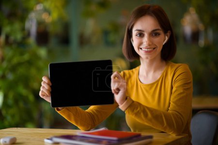 Téléchargez les photos : Cute pretty young woman with wireless earpods sitting at table, student showing digital tablet with black blank screen and smiling, recommending nice learning or business app, cafe interior, mockup - en image libre de droit