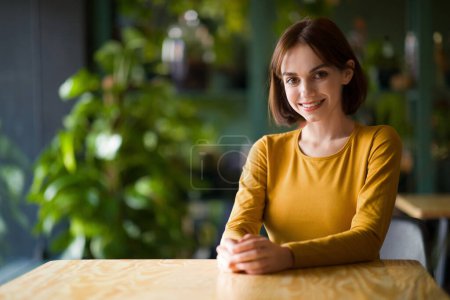 Foto de Portrait of beautiful cheerful young brunette woman with nice haircut smiling at camera while chilling at cafe, coffee shop, sitting at table, copy space. Women in business, entrepreneurship - Imagen libre de derechos