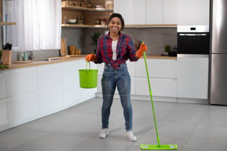 Photo for Cheerful millennial african american female in rubber gloves with bucket and mop washes floor, enjoy household chores in minimalist kitchen interior. House cleaning alone, weekend and cleaning service - Royalty Free Image