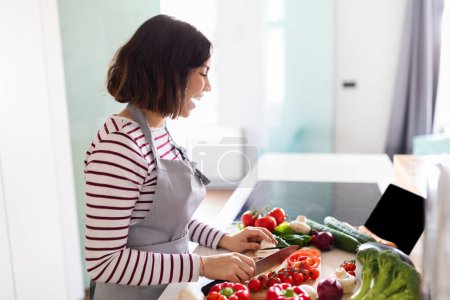 Foto de Excited pretty brunette young lady wearing grey apron watching culinary vlog and cooking at home, looking at digital pad empty screen while cutting fresh organic vegetables, copy space, side view - Imagen libre de derechos