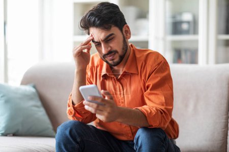 Photo for Stressed upset arab man in casual sitting on couch at home, holding cell phone, looking at gadget screen and touching his head, reading bad news, reading message, copy space - Royalty Free Image
