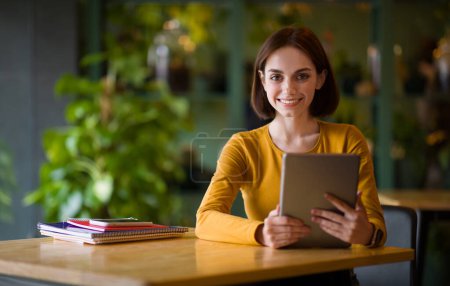Photo for Pretty cheerful young brunette woman with nice haircut sitting at table, smiling at camera, holding modern digital pad, working at coffee shop, copy space. Women in business, entrepreneurship - Royalty Free Image