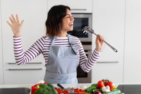 Foto de Joyful happy brunette middle eastern young woman wearing apron standing by table full of fresh organic vegetables, singing while cooking in modern kitchen, using utensil as microphone, copy space - Imagen libre de derechos